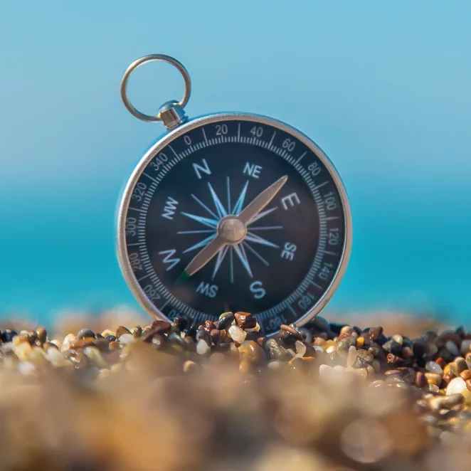 Ein klassischer Kompass steht auf einem Kieselstrand vor einem unscharfen, blauen Hintergrund (Meer oder Himmel) – Symbol für Orientierung, Strategie und Zielsetzung im Change Management bei TheMove.